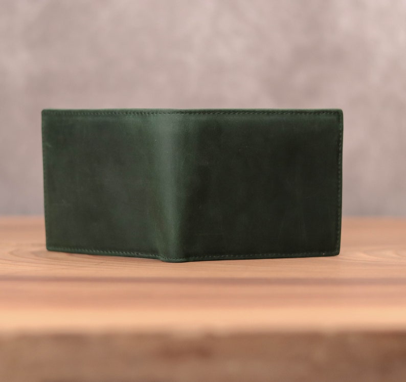 Puede incluir: Una cartera de cuero verde oscuro con un dise&ntilde;o plegado. La cartera est&aacute; sobre una superficie de madera.