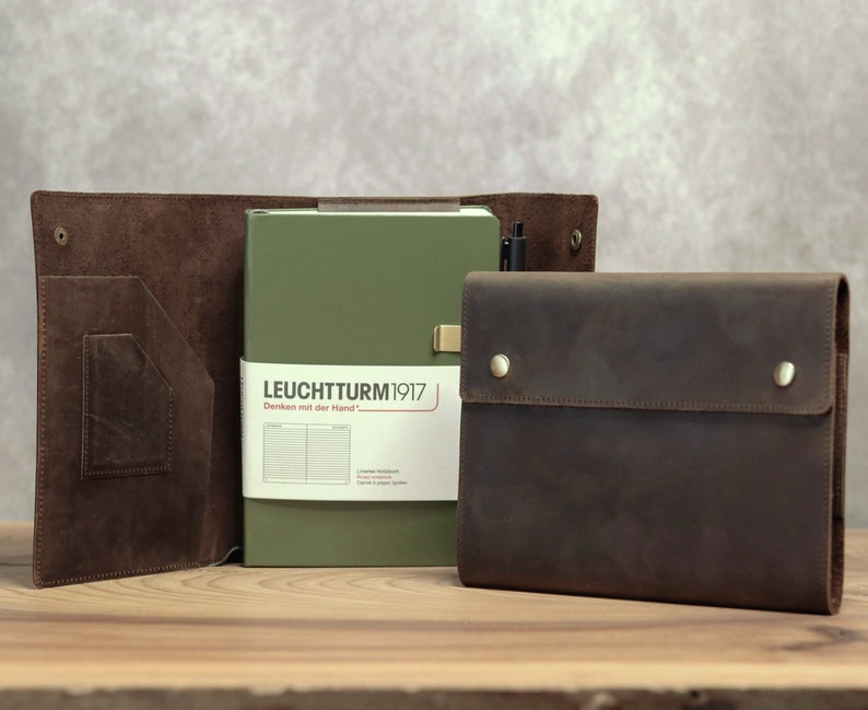 May include: A leather notebook cover and a matching dark brown leather folio with a green notebook inside. The notebook has the text "LEUCHTTURM1917" on the cover. A black pen is in the notebook.
