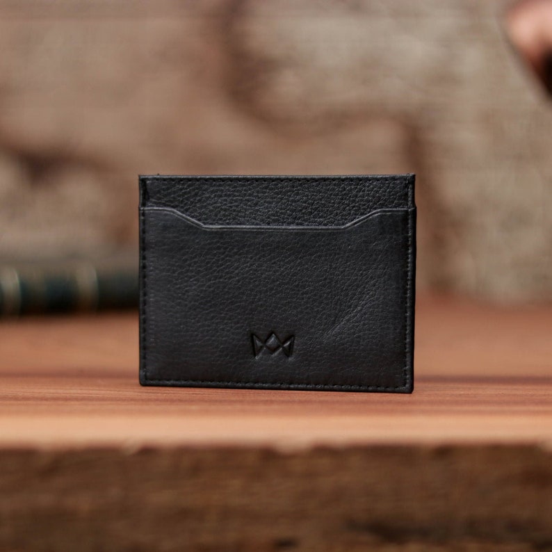May include: A black leather card holder with a subtle textured finish. The card holder has a single pocket and a small logo on the front.