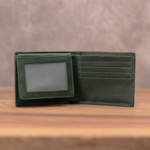Puede incluir: Una cartera de cuero verde oscuro con m&uacute;ltiples ranuras para tarjetas y una ventana de identificaci&oacute;n transparente. La cartera est&aacute; abierta y sobre una superficie de madera.