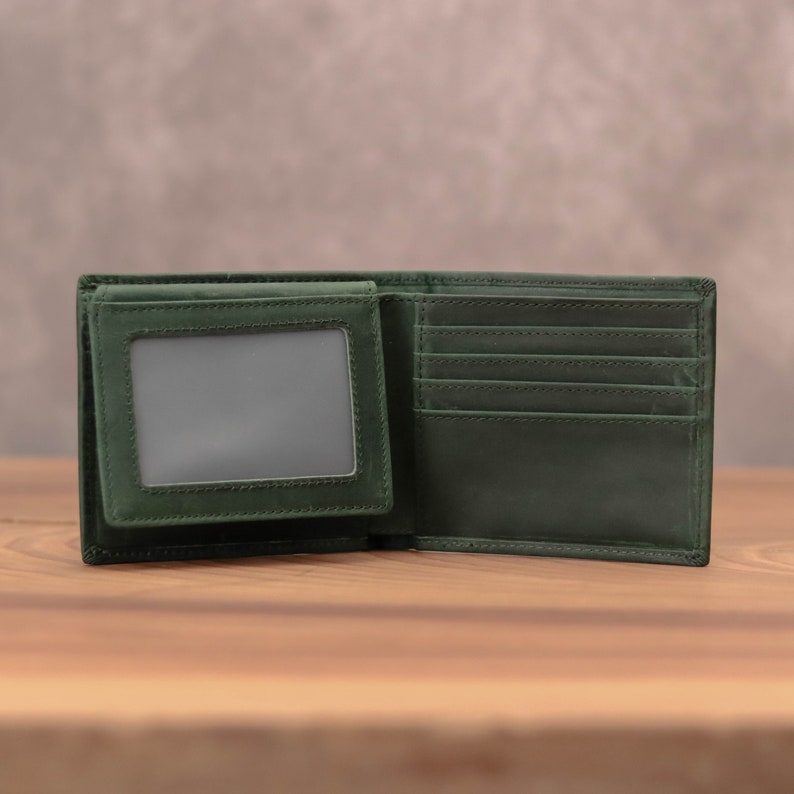Puede incluir: Una cartera de cuero verde oscuro con m&uacute;ltiples ranuras para tarjetas y una ventana de identificaci&oacute;n transparente. La cartera est&aacute; abierta y sobre una superficie de madera.