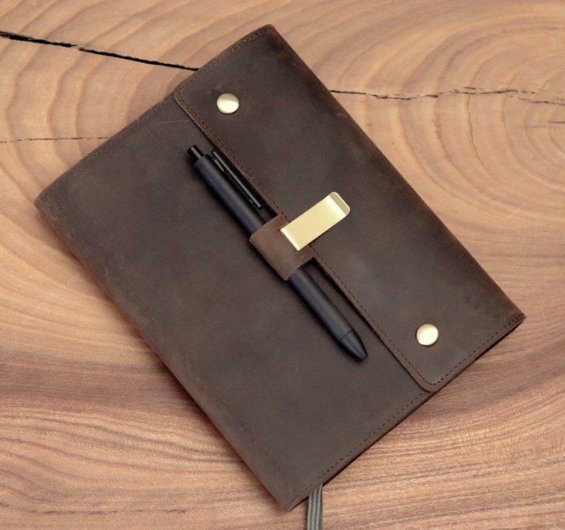 May include: A brown leather journal with a pen holder and a black pen. The journal has a flap closure with two brass-colored button studs. A gold-colored metal clip secures the pen. The journal is on a wooden surface.