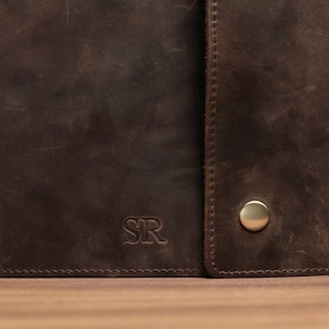 May include: Close-up of a dark brown leather wallet with a snap closure. The wallet has a distressed texture and the initials "SR" are embossed on the front. A metal snap is visible on the right side.
