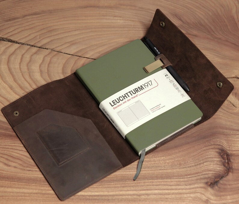 May include: An olive green notebook with the text "LEUCHTTURM1917" is secured by a white band and a pen holder. The notebook is inside a brown leather cover with a pocket and snap closure. The cover is open on a wooden surface.