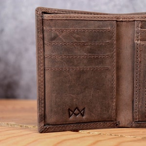 May include: Open brown leather wallet with multiple card slots and a bill compartment. The wallet has a distressed finish and a stitched border. A small emblem is embossed on the lower right corner.