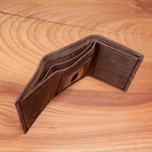 May include: Open brown leather wallet with multiple card slots and a clear ID window. The wallet is displayed open, revealing the interior compartments and the texture of the leather. The wallet is on a wooden surface.