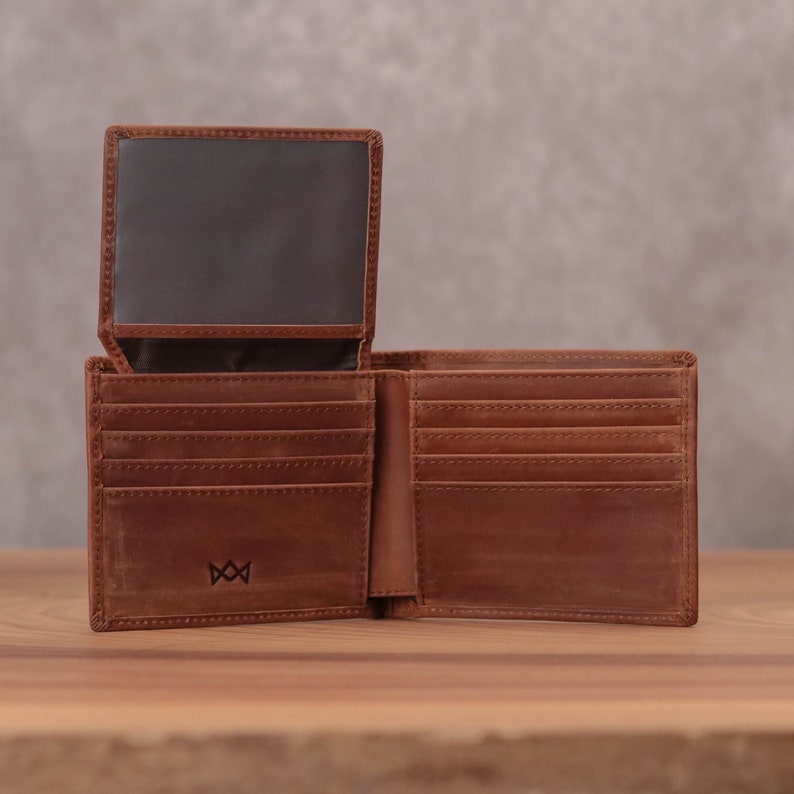 Puede incluir: Una cartera de cuero marr&oacute;n con m&uacute;ltiples ranuras para tarjetas y un portafolios. La cartera est&aacute; abierta y el interior es visible. La cartera tiene un peque&ntilde;o logotipo en la parte delantera.