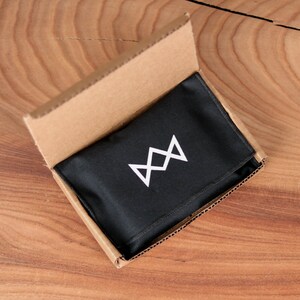 May include: An open cardboard box with a black fabric pouch inside. The pouch has a white logo with a stylized crown design.