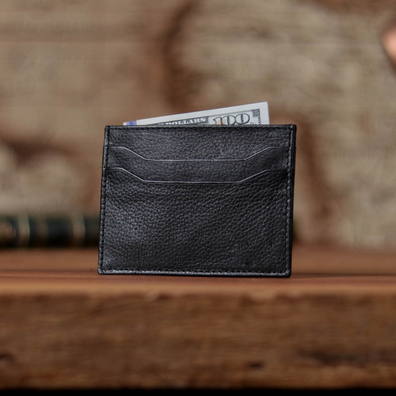 May include: A black leather card holder with multiple slots for cards. A dollar bill is partially visible inside the holder.