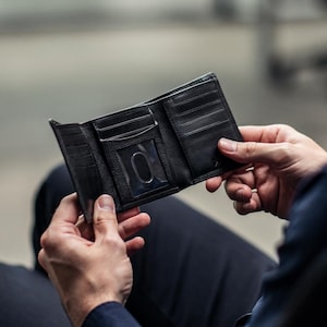 May include: Open black leather trifold wallet with multiple card slots and an ID window. The wallet is held open, showcasing its interior design. The leather appears textured, and the wallet is empty.