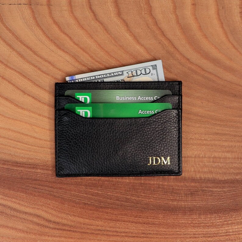 May include: Black leather card holder with two green credit cards and a $100 bill inside. The card holder has the initials "JDM" embossed in gold on the front.