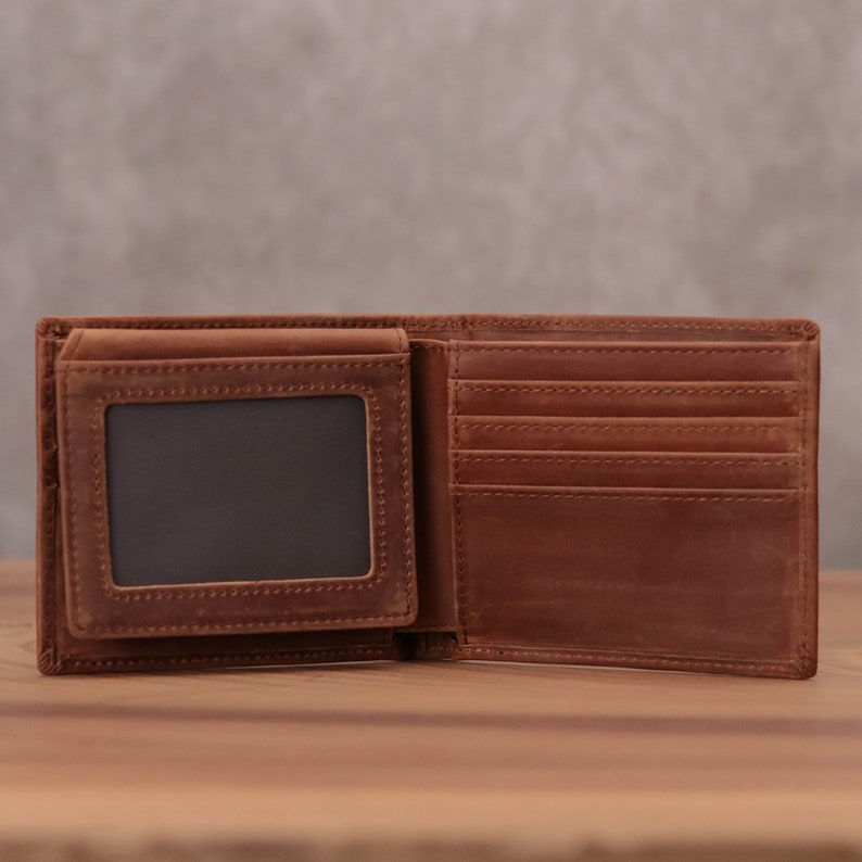 Puede incluir: Una cartera de cuero marr&oacute;n con una ventana de pl&aacute;stico transparente para una tarjeta de identificaci&oacute;n. La cartera est&aacute; abierta y tiene m&uacute;ltiples ranuras para tarjetas y un portafolios.