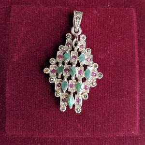 Può includere: Un ciondolo d'argento con un design geometrico con pietre preziose verdi e rosse. Il ciondolo è posto su uno sfondo di velluto rosso.
