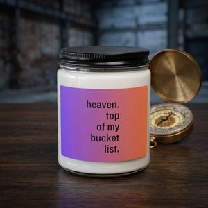 Pode incluir: Uma vela branca num frasco de vidro transparente com tampa preta. O rótulo apresenta um gradiente de roxo para laranja com o texto preto: heaven. top of my bucket list. Uma bússola vintage cor de latão está aberta ao lado do frasco.