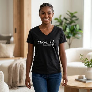 May include: Black V-neck t-shirt featuring the words "even if" and the Bible verse Daniel 3:18 in white script. The shirt is paired with blue jeans. The person is smiling.