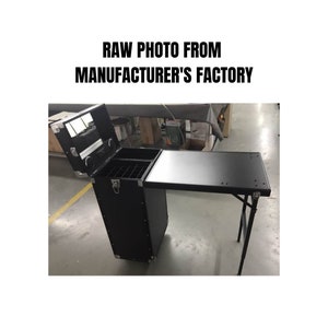 May include: Black rolling makeup case with a fold-out table. The case has a top compartment with a mirror and storage, a middle section with dividers, and a lower storage area. The text "RAW PHOTO FROM MANUFACTURER'S FACTORY" is at the top.