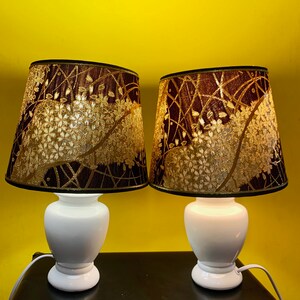 May include: Two white table lamps with brown and gold patterned lampshades. The lampshades have a floral design with gold accents.