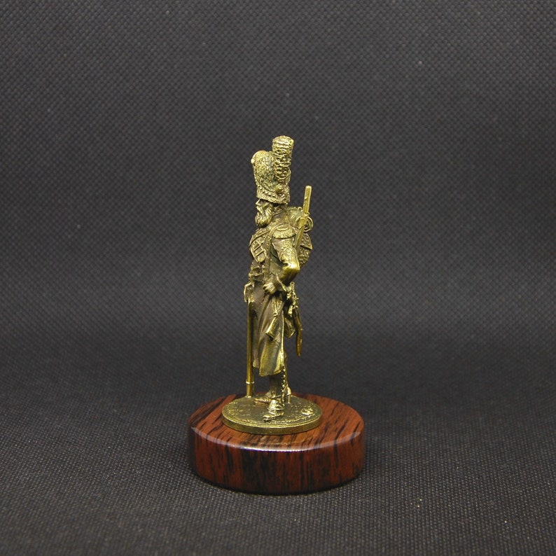 May include: A gold-colored metal figurine of a soldier wearing a tall, feathered hat and holding a rifle. The figurine is standing on a wooden base.