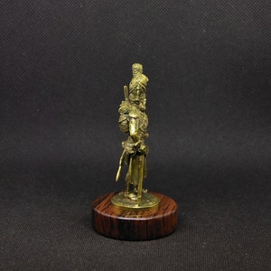 May include: A bronze figurine of a soldier wearing a plumed helmet and holding a sword. The figurine is standing on a wooden base.