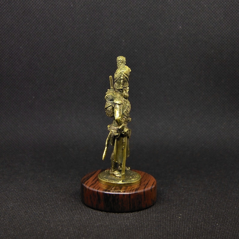May include: A bronze figurine of a soldier wearing a plumed helmet and holding a sword. The figurine is standing on a wooden base.