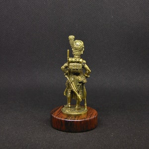 May include: A gold-colored figurine of a soldier wearing a tall, feathered hat and holding a rifle. The figurine is standing on a wooden base.