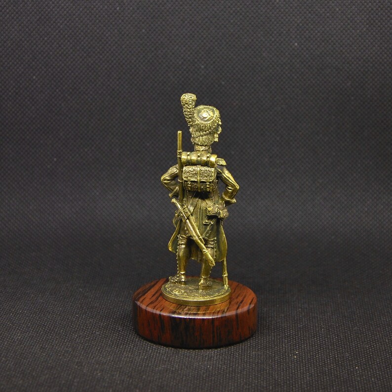 May include: A gold-colored figurine of a soldier wearing a tall, feathered hat and holding a rifle. The figurine is standing on a wooden base.