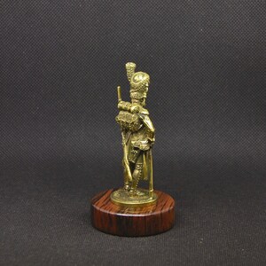 May include: A gold-colored miniature figurine of a soldier standing on a wooden base. The soldier is wearing a tall, feathered hat and holding a rifle.