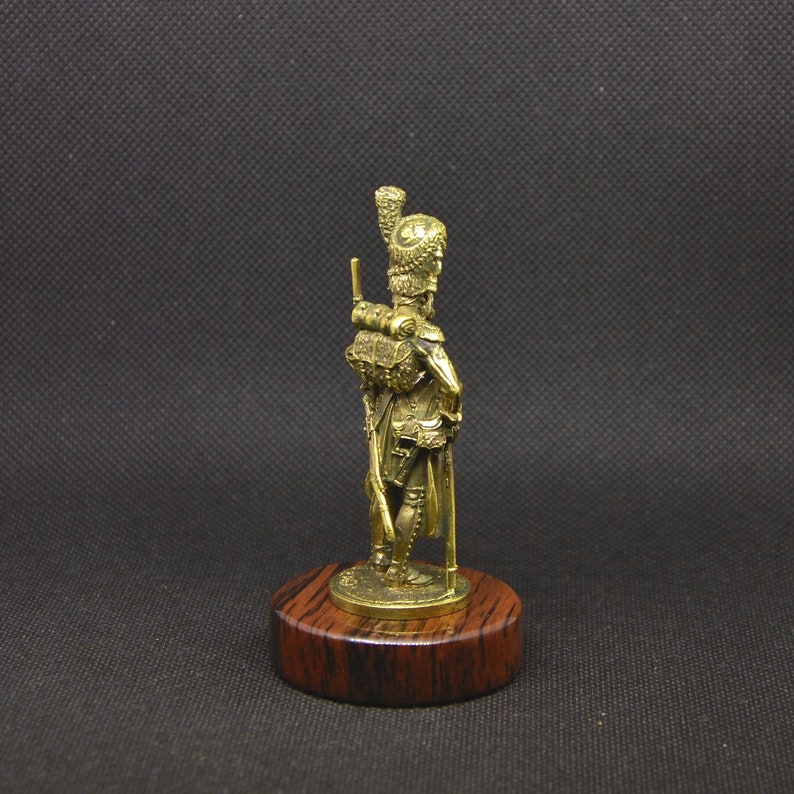 May include: A gold-colored miniature figurine of a soldier standing on a wooden base. The soldier is wearing a tall, feathered hat and holding a rifle.