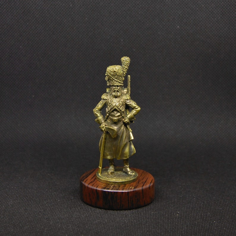 May include: A bronze figurine of a soldier wearing a tall, feathered hat and holding a musket. The figurine is standing on a wooden base.
