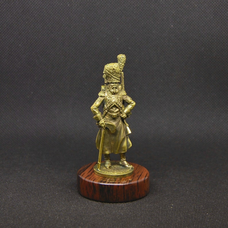 May include: A bronze figurine of a soldier in a military uniform, standing on a wooden base. The soldier is wearing a tall hat with a plume, and is holding a rifle in his right hand.