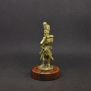 May include: A gold-colored figurine of a soldier wearing a tall, feathered hat and carrying a rifle. The figurine is standing on a wooden base.