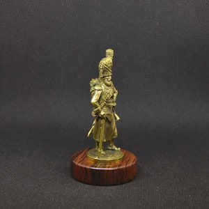 May include: A gold-colored metal figurine of a soldier wearing a tall, feathered hat and holding a rifle. The figurine is standing on a wooden base.