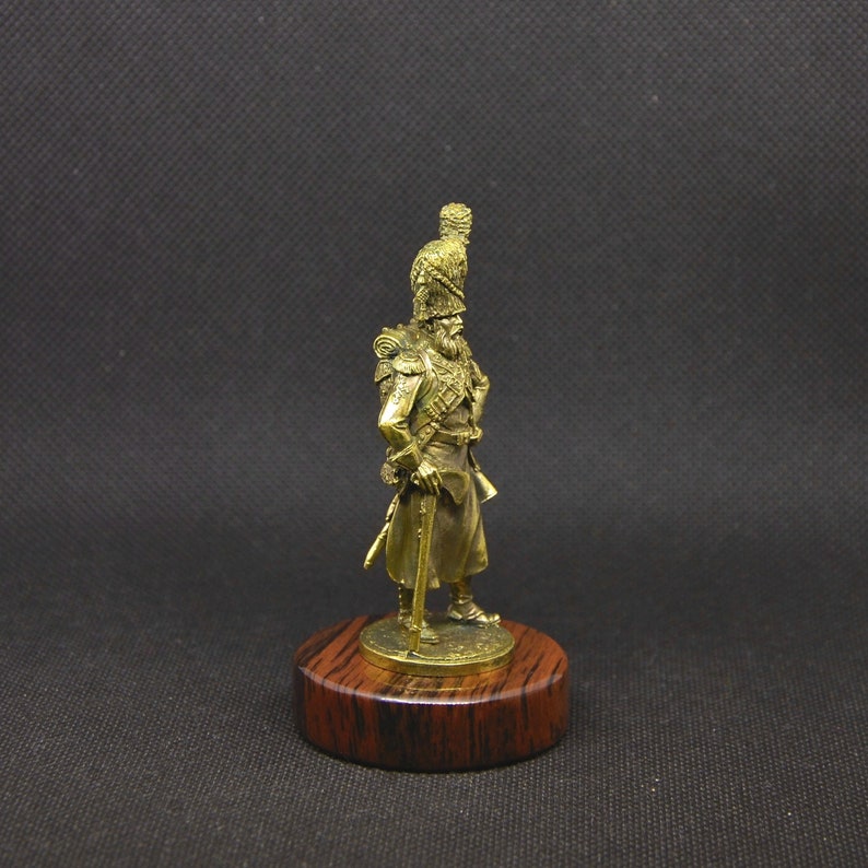 May include: A gold-colored metal figurine of a soldier wearing a tall, feathered hat and holding a rifle. The figurine is standing on a wooden base.
