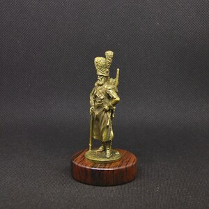 May include: A gold-colored metal figurine of a soldier wearing a tall, ornate hat and holding a rifle. The figurine is standing on a wooden base.