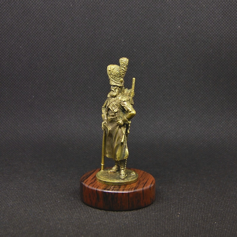 May include: A gold-colored metal figurine of a soldier wearing a tall, ornate hat and holding a rifle. The figurine is standing on a wooden base.