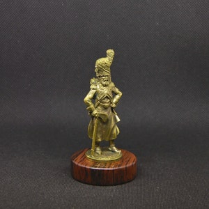 May include: A gold-colored figurine of a soldier wearing a tall, feathered hat and holding a staff. The figurine is standing on a wooden base.