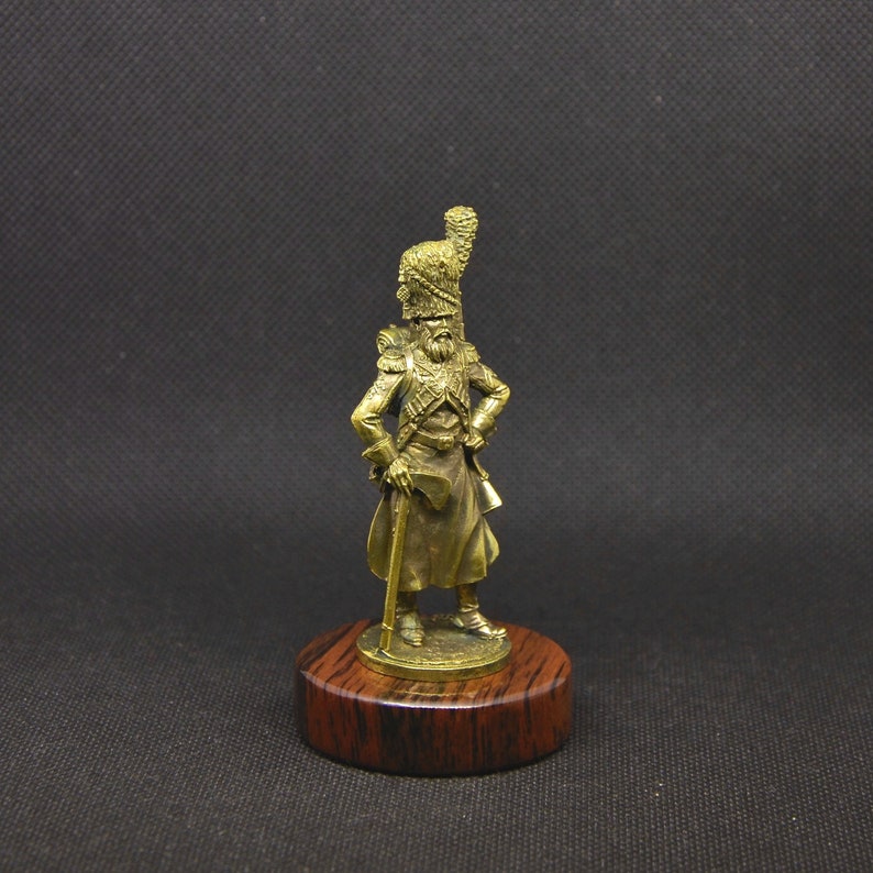 May include: A gold-colored figurine of a soldier wearing a tall, feathered hat and holding a staff. The figurine is standing on a wooden base.