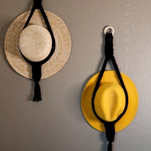 May include: Two hats hanging on a wall using macrame rope hangers. The hat on the left is a straw hat with a brown band. The hat on the right is a yellow felt hat with a black band.