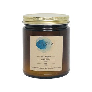 Puede incluir: Una vela de vidrio marrón en un frasco con tapa dorada. La etiqueta dice "B.B.SHA" y "Warm & Sweet" con la descripción "Rum & Honey". La vela es de 7 oz (198,447 g) y es una vela de soja y coco hecha en Florida.