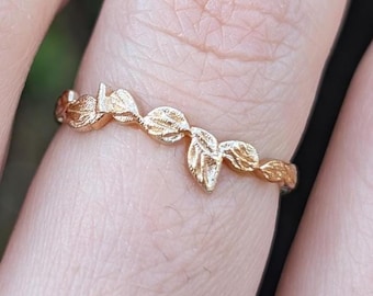Puede incluir: Un anillo dorado con un delicado diseño de hojas. El anillo tiene una banda fina y está adornado con varias hojas pequeñas y detalladas. Las hojas están dispuestas en un patrón repetitivo alrededor de la circunferencia del anillo.