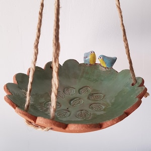 Può includere: Una ciotola in ceramica sospesa con bordo smerlato e interno smaltato verde con motivi a foglia in rilievo. Due piccoli uccelli in ceramica colorati, con petto giallo e ali blu, sono appollaiati sul bordo. Sospesa tramite una corda di iuta.