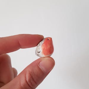May include: A small, ceramic robin figurine with a red breast and a gray and white wing design. The bird is held between fingers against a white background. The figurine is approximately 1 inch in height.