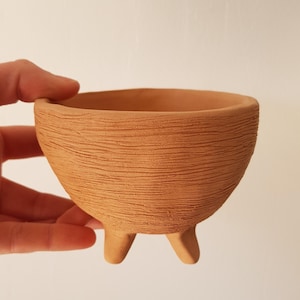 Può includere: Una piccola ciotola color terracotta con una superficie strutturata a righe orizzontali. La ciotola ha tre gambe corte e una forma arrotondata. La ciotola è tenuta da una mano.