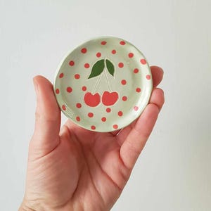 Op de afbeelding: Kleine, ronde keramische schaal met een lichtgroene achtergrond en rode stippen. In het midden een paar rode kersen met groene bladeren. De schaal wordt in een hand gehouden.