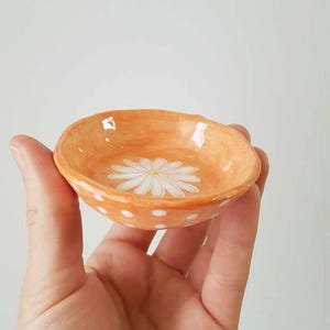 Op de afbeelding: Een klein, oranje keramiek schaaltje met witte stippen en een witte, geschilderde madeliefje in het midden.