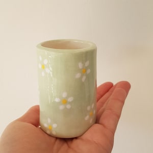 Peut inclure: Un petit vase cylindrique en céramique de couleur vert clair, orné de motifs floraux blancs en forme de marguerites avec des centres jaunes. Le vase est tenu dans une main, mettant en valeur sa taille et son design. Le vase mesure environ 7.6 cm de haut.
