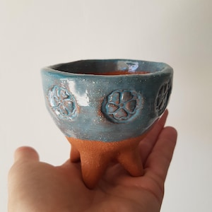 Könnte beinhalten: Eine kleine, dreibeinige Keramikschale mit einem Terrakotta-Sockel und einem glänzenden, gesprenkelten blauen Oberteil. Der blaue Bereich zeigt ein sich wiederholendes, geprägtes Blumenmuster an der Außenseite, während das Innere aus unglasiertem Terrakotta besteht.