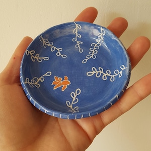 May include: A small, round, blue ceramic dish with white leaf and vine designs. An orange floral design is in the centre. The rim has a white striped pattern. The dish is held in a hand.