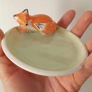 May include: A ceramic trinket dish with a sleeping fox figurine. The dish is oval-shaped with a light green interior and a white rim. The fox is orange and white with black accents.