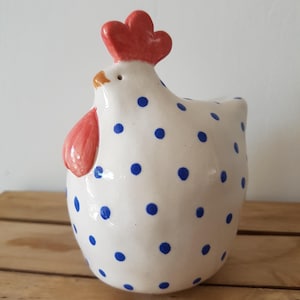 Può includere: Una statuetta di gallina in ceramica con base bianca e un motivo a piccoli pois blu. La gallina presenta una cresta rosso brillante sulla parte superiore della testa e un bargiglio rosso sotto il becco, con un piccolo becco marrone.